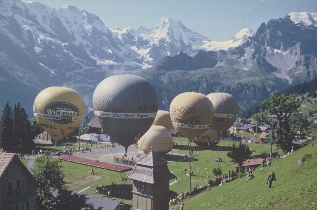 Aufstieg von sieben Ballons bei der 4. Internationalen Ballonsportwoche in Mürren (Kanton Bern) am 19. August 1965. Das auf 1650 m gelegene Dorf Mürren war der ideale Startplatz für Hochalpen-Ballonaufstiege. Im Wettbewerb wurden nicht Dauer, Höhe oder zurückgelegte Distanz berück- sichtigt, sondern die sichere Landung und der schriftliche Fahrtbericht mit eingezeichneter Flugstrecke, die von der Jury bewertet wurden. Signatur: Staatsarchiv des Kantons Zürich, Ballongruppe Zürich, Z 1037.265.2.